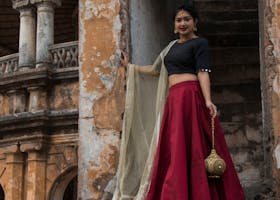A woman in a vibrant red skirt and black blouse stands gracefully on rustic stone stairs in an ancient Indian setting.